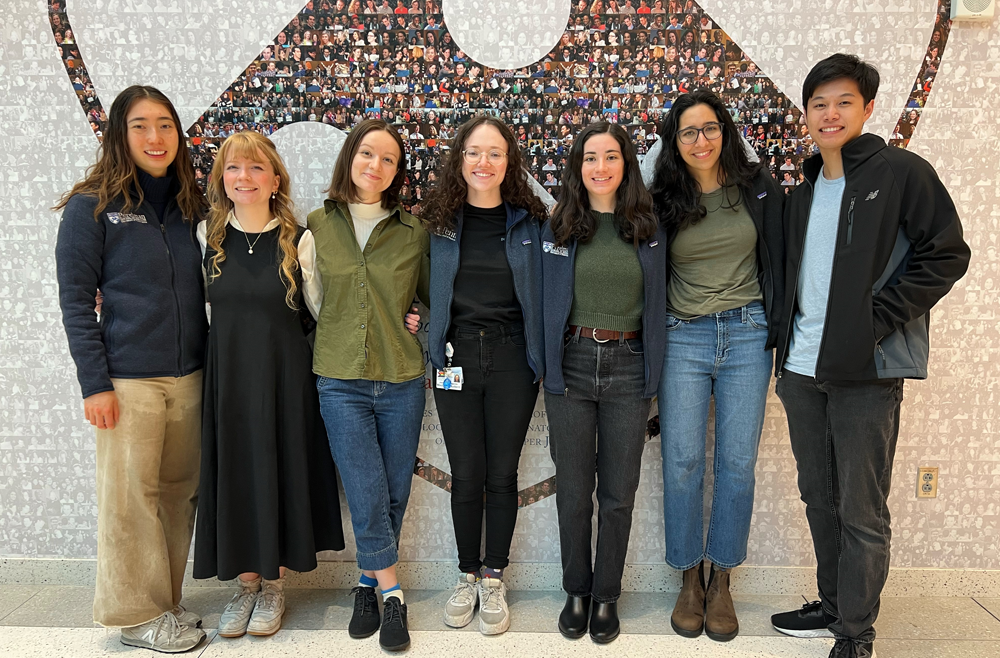 Medical students Catherine Shen, Elizabeth Whidden, Phoebe Cunningham, Jess Campanile, Sarah Wornow, Isha Thapar, Jonathan Szeto stand in front of a mural of the Penn shield.